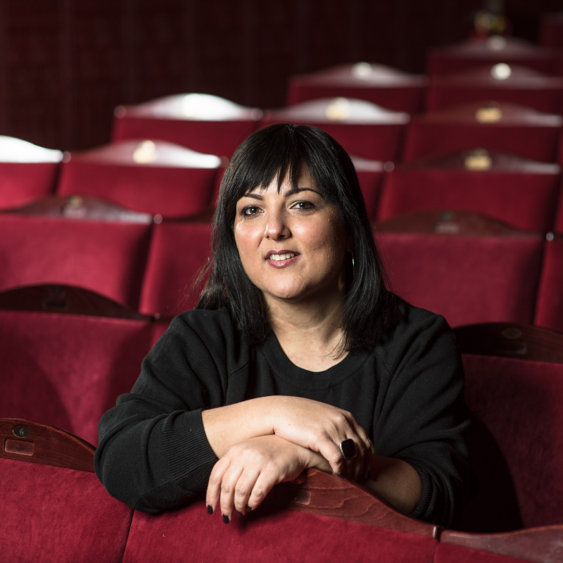 Artistic Director, Nadia Fall, sits in the auditorium amongst rows of red velvet covered chairs. She leans forward, hands resting on the top of the chairs in front of her.